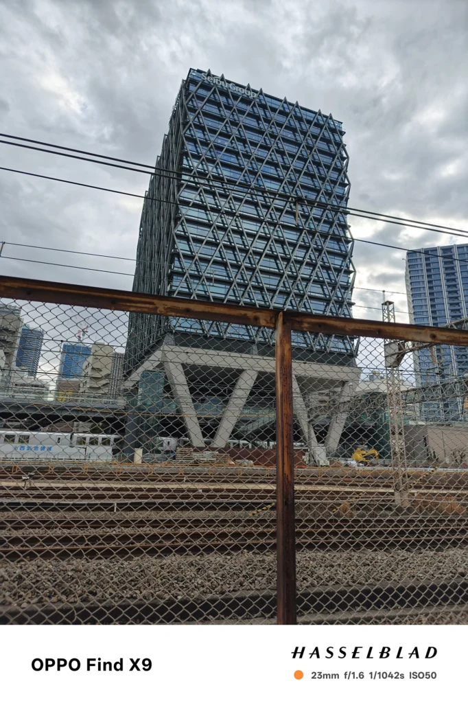 端末名：OPPO Find X9／カメラ：メイン（広角）／焦点距離：23mm相当／内容：線路脇の金網フェンス越しに、格子外装の高層ビルと架線が交差する曇天の都市景観