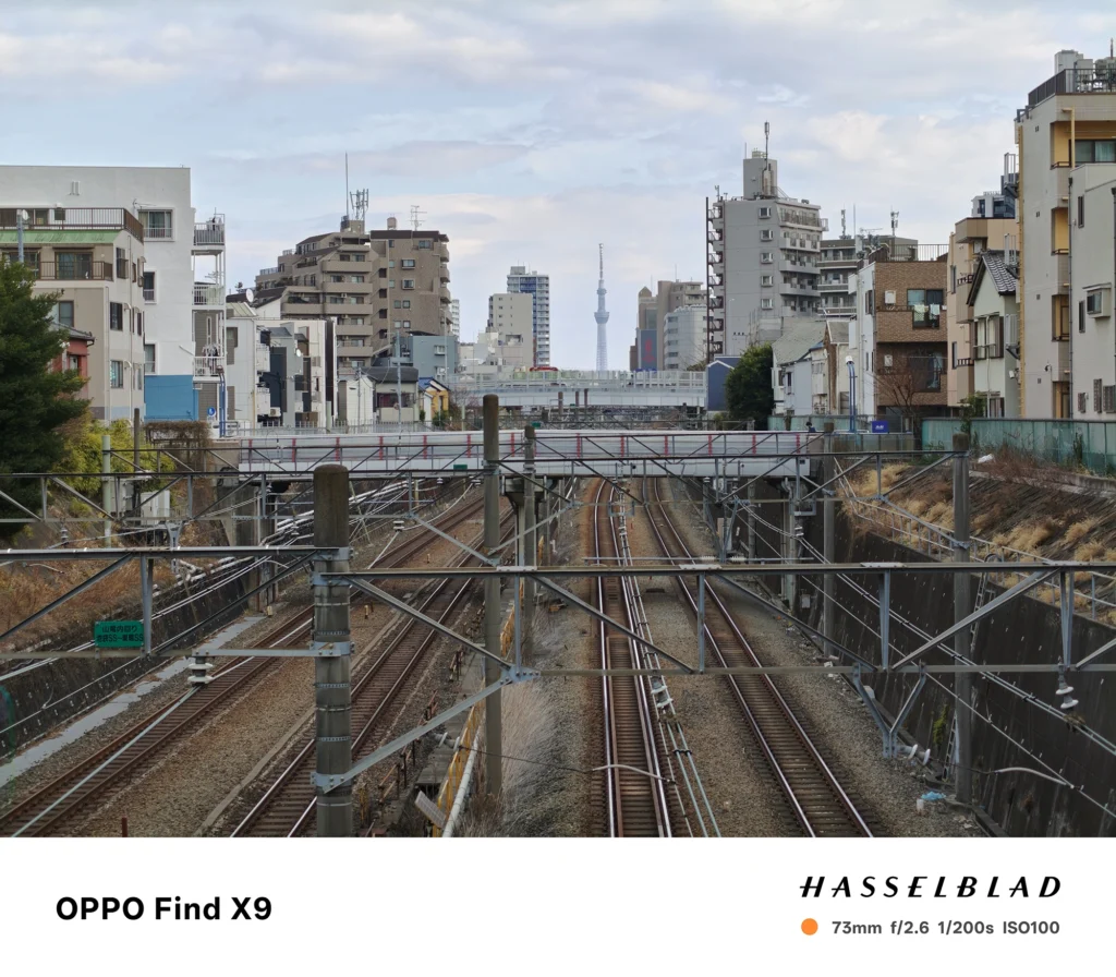 端末名：Find X9／カメラ：望遠／焦点距離：確認不能（mm相当）mm相当／内容：複数の線路と架線を遠方から引き寄せて圧縮感を出し、屋外の昼光で街並みと線路の直線を整理して捉える