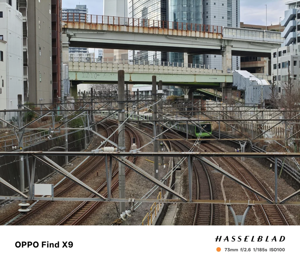 Find X9／カメラ：望遠／焦点距離：確認不能（mm相当）mm相当／内容：カーブする線路と走行中の列車を引き寄せて背景を圧縮し、屋外の昼光で架線や高架の構造物を細密に描写