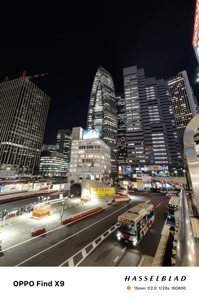 Find X9／カメラ：超広角／焦点距離：15mm相当／内容：工事柵やバスの走る交差点を広く捉え、強い照明と車灯が混在する夜の都市風景