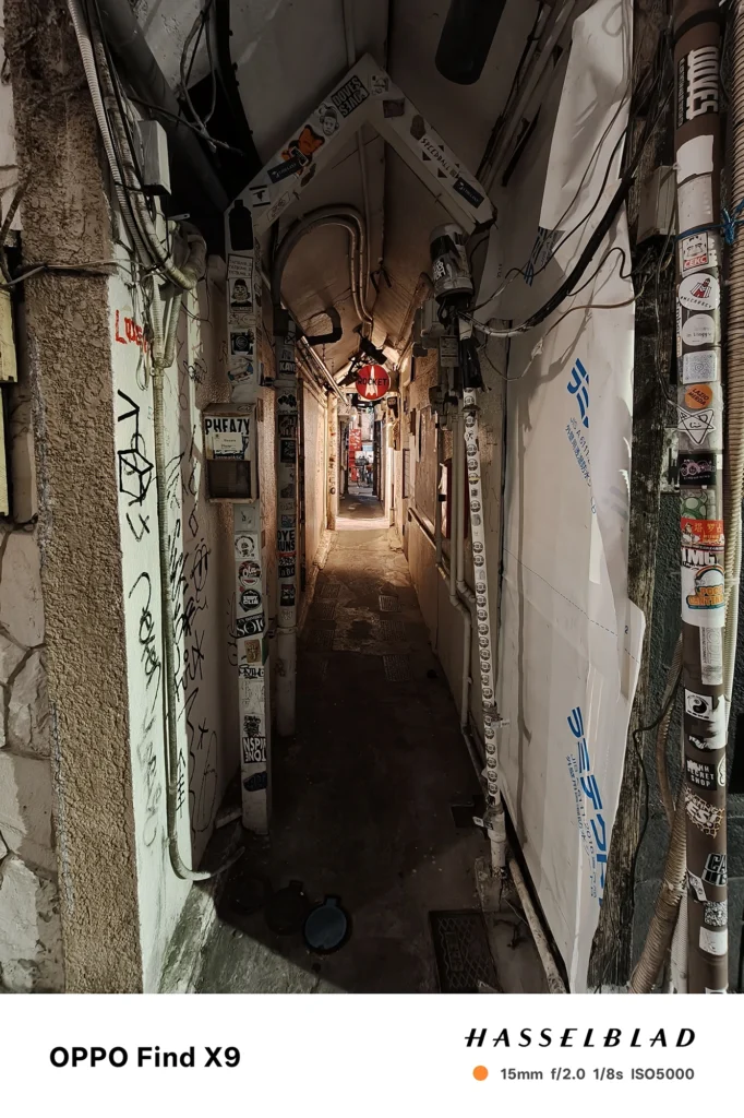 端末名：Find X9／カメラ：超広角／焦点距離：15mm相当／内容：ステッカーや配管が密集した細い路地を写し、壁面照明が点在する夜の路地風景