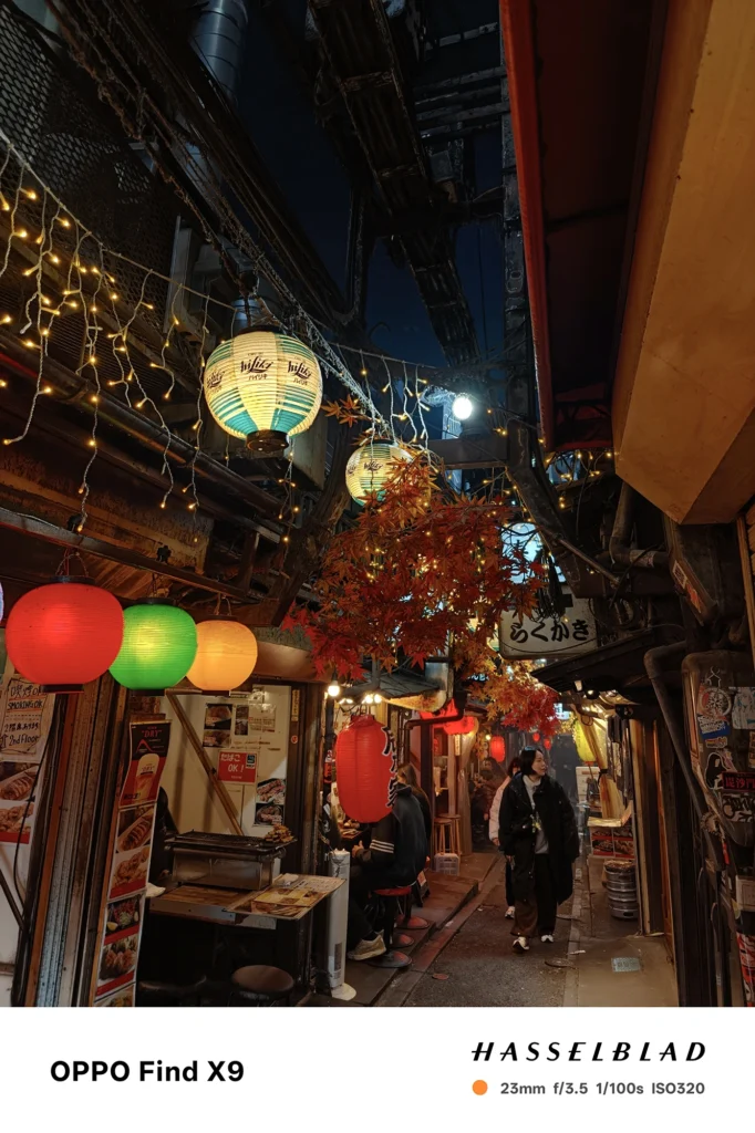 Find X9／カメラ：メイン（広角）／焦点距離：確認不能（mm相当）mm相当／内容：提灯と串焼き店が並ぶ夜の細い飲み屋街を捉え、暖色照明とネオンが人の往来を照らす路地の雰囲気