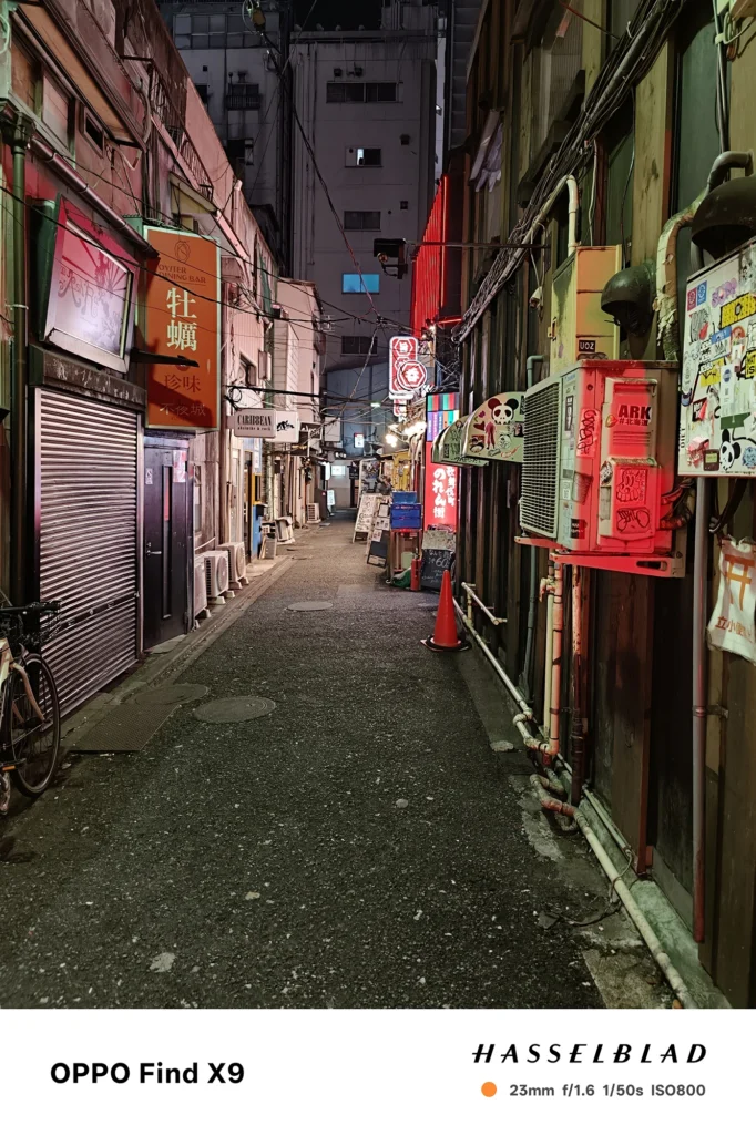 Find X9／カメラ：メイン（広角）／焦点距離：確認不能（mm相当）mm相当／内容：看板と街灯が並ぶ夜の路地を奥行き方向に写し、配線や店舗外観の細部が照明に浮かぶ落ち着いた雰囲気