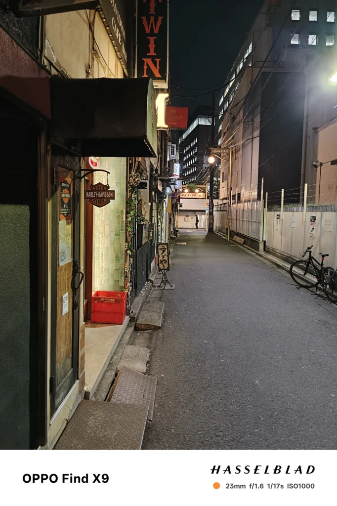 Find X9／カメラ：メイン（広角）／焦点距離：確認不能（mm相当）mm相当／内容：夜の裏通りを斜めに見通し、店舗看板と街灯が路面を照らし自転車が停まる静かな路地の雰囲気