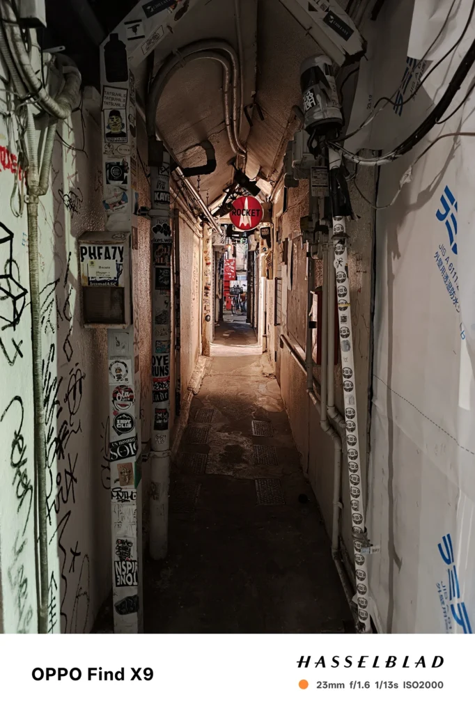 Find X9／カメラ：メイン（広角）／焦点距離：確認不能（mm相当）mm相当／内容：ステッカーと配管が並ぶ夜の細い通路を奥まで写し、天井灯の暖色光が壁面の質感を浮かび上がらせる