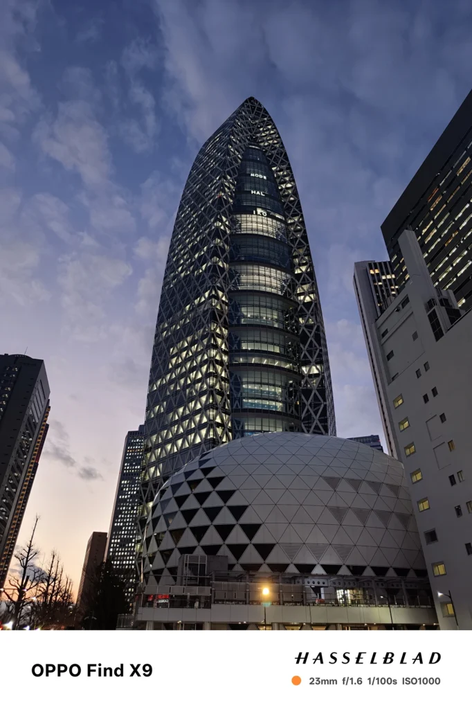 端末名：Find X9／カメラ：メイン（広角）／焦点距離：確認不能（mm相当）mm相当／内容：夕暮れの高層ビル群を見上げ、窓明かりと外装パターンが浮かぶ夜景を空のグラデーションと街灯で描写