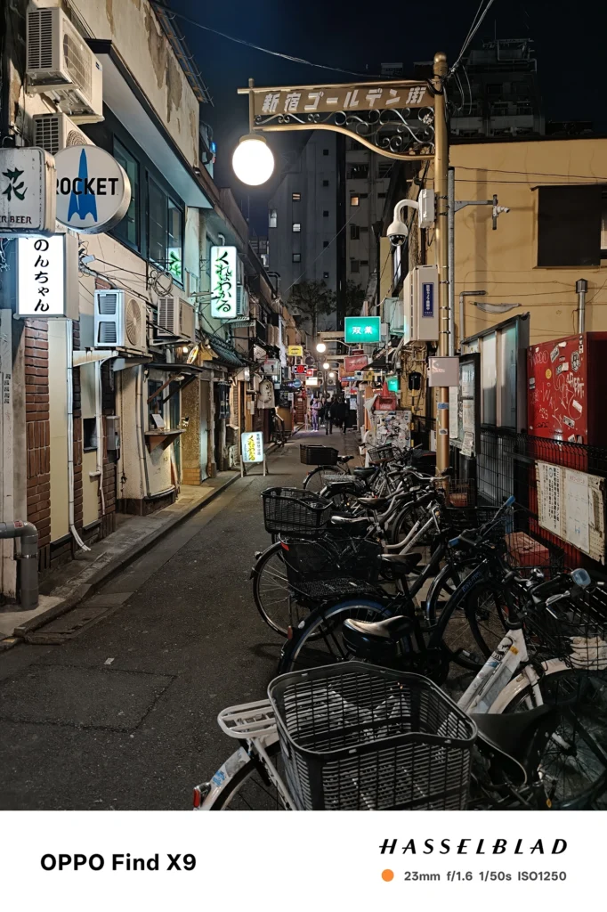 端末名：Find X9／カメラ：メイン（広角）／焦点距離：確認不能（mm相当）mm相当／内容：自転車が並ぶ夜の飲み屋街路地を正面に捉え、街灯と看板の光が狭い通りの奥行きを強調する
