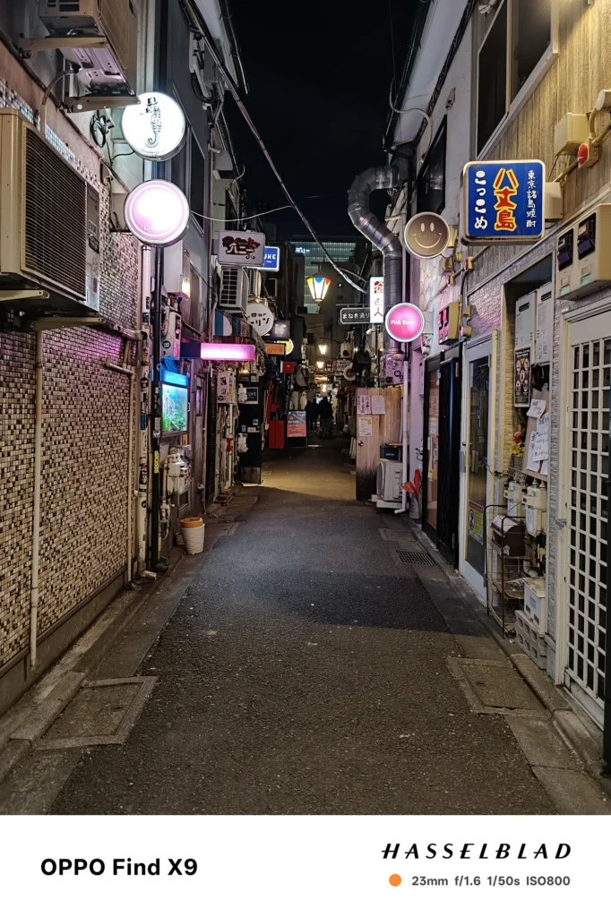 Find X9／カメラ：メイン（広角）／焦点距離：確認不能（mm相当）mm相当／内容：ピンクや白のネオンが並ぶ夜の路地を正面に捉え、看板光と街灯が壁面や配線を照らす都会の裏通り