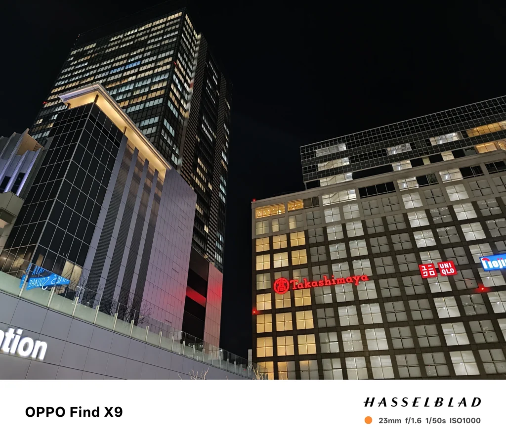 Find X9／カメラ：メイン（広角）／焦点距離：確認不能（mm相当）mm相当／内容：夜のビル群を見上げ、窓明かりと企業ロゴが暗い空に浮かぶ都会夜景を白色照明の輪郭で引き締める