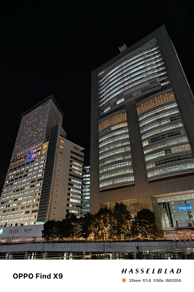 Find X9／カメラ：メイン（広角）／焦点距離：確認不能（mm相当）mm相当／内容：高層ビルを見上げた夜景を捉え、窓明かりの反復と外装ラインが街灯の光で強調される静かな都会の雰囲気