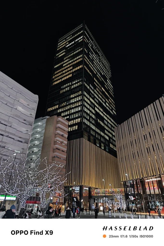 端末名：Find X9／カメラ：メイン（広角）／焦点距離：確認不能（mm相当）mm相当／内容：イルミネーションの木々と広場を前景に高層ビルを見上げ、街灯と窓明かりが暗い空に浮かぶ冬の夜景