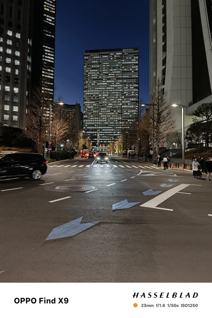 端末名：Find X9／カメラ：メイン（広角）／焦点距離：確認不能（mm相当）mm相当／内容：高層ビルに挟まれた夜の大通りを正面から捉え、車のライトと街灯が路面反射を作る都市夜景