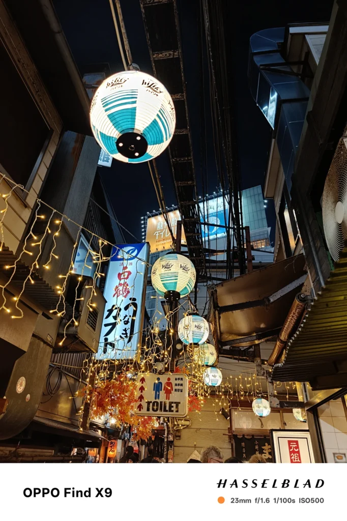 端末名：Find X9／カメラ：メイン（広角）ポートレート／焦点距離：23mm相当／内容：飲食店の提灯と看板を手前に、路地の人混みと灯りを背景ボケでまとめた夜景ポートレート