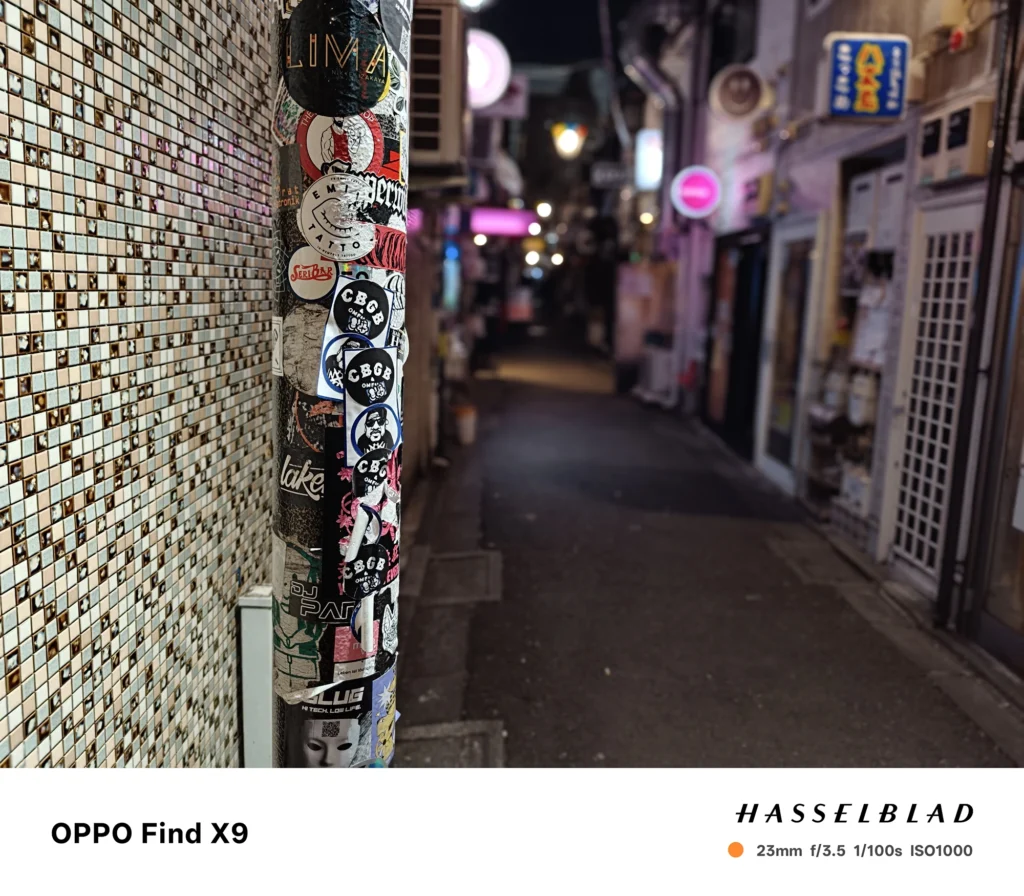 端末名：Find X9／カメラ：メイン（広角）ポートレート／焦点距離：23mm相当／内容：ステッカーの貼られた電柱を主役に、路地のネオンや灯りを背景ボケで表現した夜景ポートレート