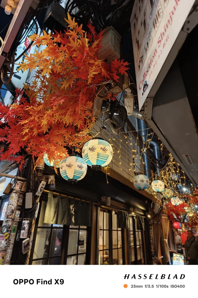 端末名：Find X9／カメラ：メイン（広角）ポートレート／焦点距離：23mm相当／内容：ステッカーの多い電柱を前景に、路地奥の紫系ネオンや街灯を背景ボケにした夜景ポートレート