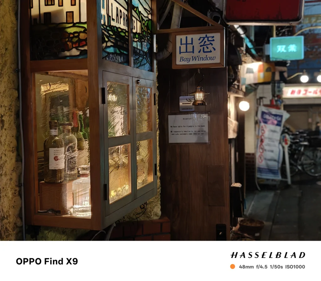 バー入口の看板とディスプレイを48mmで撮影（OPPO Find X9）。木材とガラスの質感、背景ボケを描写。