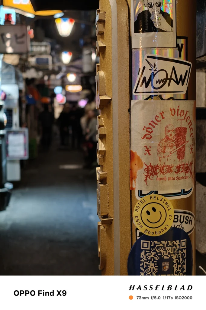 ステッカーだらけの柱を73mmで撮影（OPPO Find X9）。近接の解像と奥の路地の大きなボケを描写。