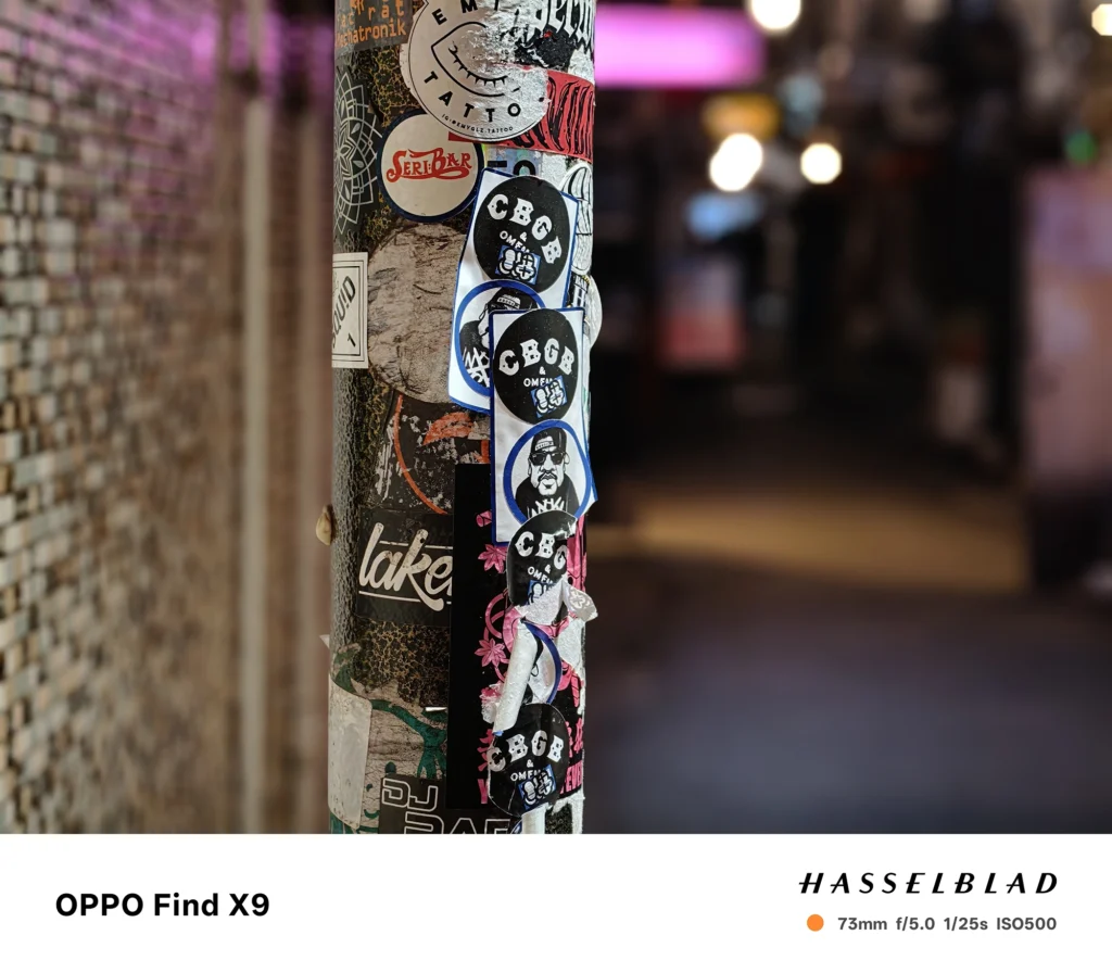 夜の路地でステッカーの柱を73mmポートレート撮影（OPPO Find X9）。背景のネオン光を柔らかくぼかした作例。