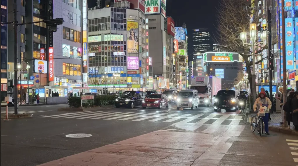 端末名：OPPO Find X9／カメラ：確認不能／焦点距離：確認不能mm相当／内容：夜の繁華街交差点で走行する車のヘッドライトとネオン看板、歩道の人影を広く捉えたナイトストリートビュー