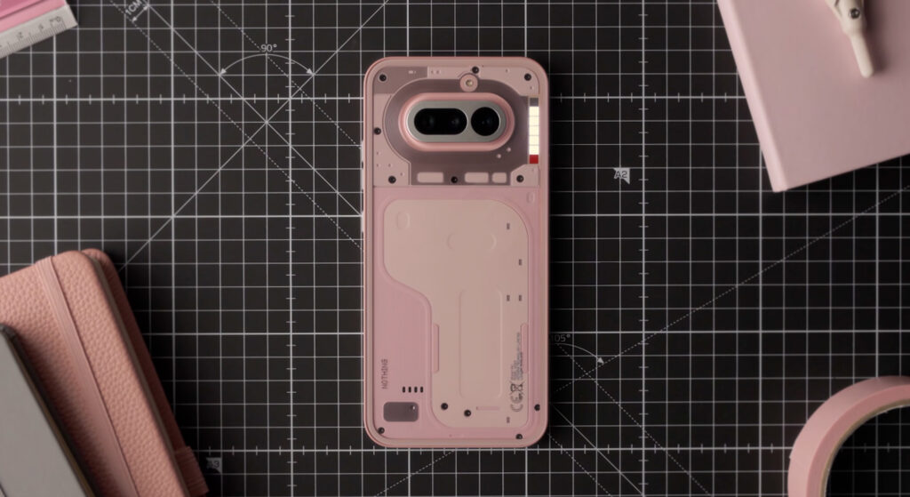 Nothing Phone (4a) ピンクモデル。カッティングマット上に置かれ、透明な背面に透過する内部パーツのトーンが調和している。