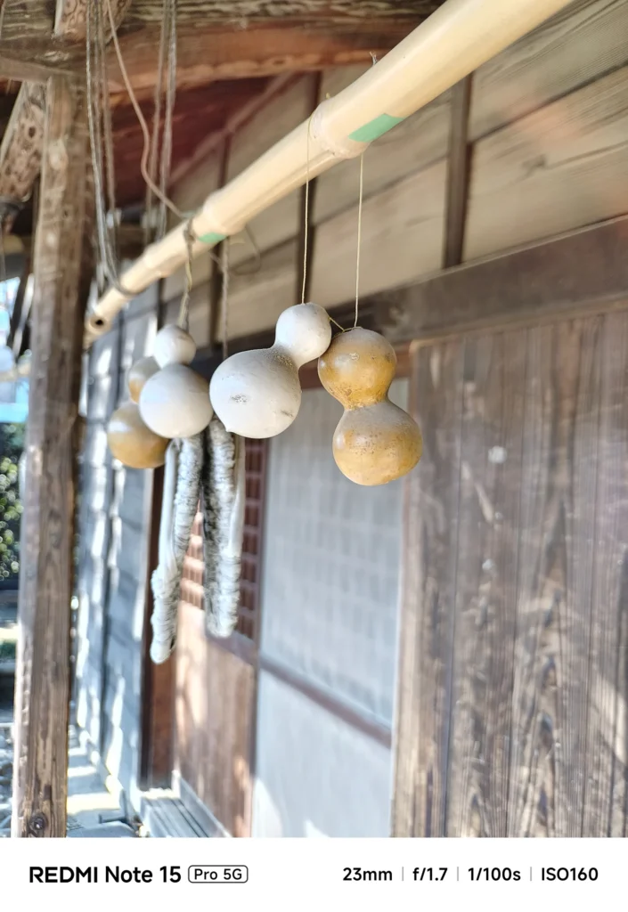 Redmi Note 15 Pro 5G＋外付け望遠（約40mm相当）で撮影した吊るされた瓢箪のクローズアップ、木壁を大きくぼかして主題を強調