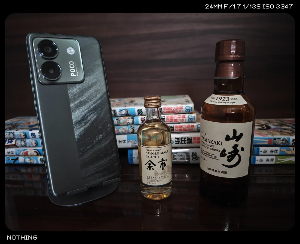 Nothing Phone 3で暗めの屋内に並ぶスマホ、ウイスキーボトル、漫画本を等倍撮影した作例