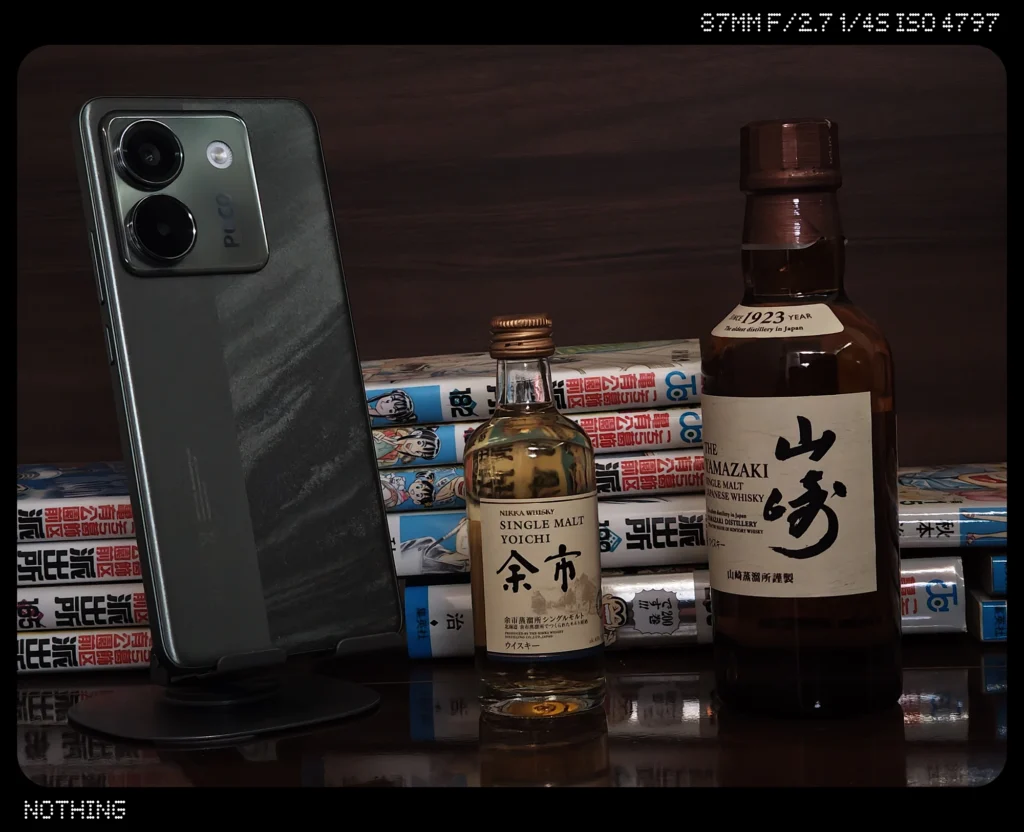 Nothing Phone 3で暗めの屋内に並ぶスマホ、ウイスキーボトル、漫画本をズーム撮影した作例