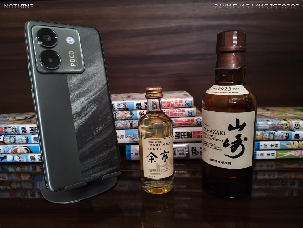 Nothing Phone 3aで暗い木目背景のスマホ、ウイスキーボトル、漫画本を等倍撮影した作例
