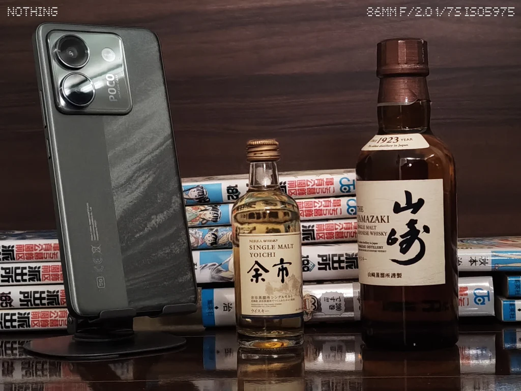 Nothing Phone 3aで暗めの屋内にあるスマホ、ウイスキーボトル、漫画本をズーム撮影した作例