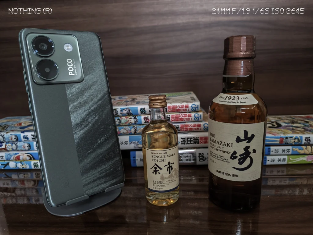 Nothing Phone 3a Liteで暗めの屋内に置いたスマホ、ウイスキーボトル、漫画本を等倍撮影した作例
