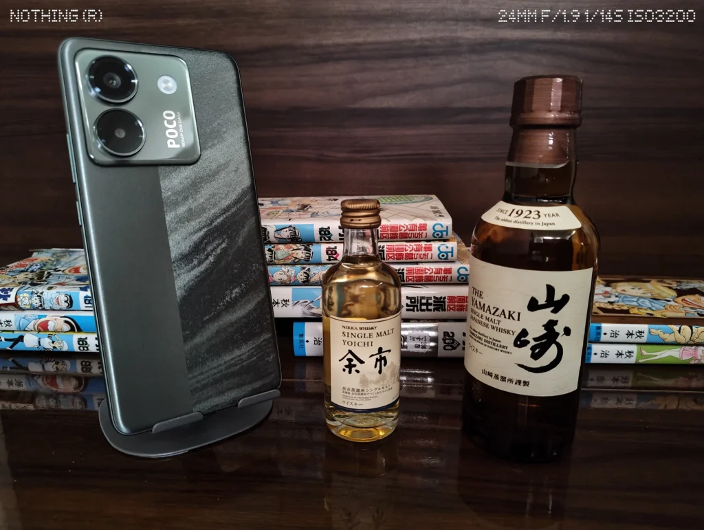 Nothing Phone 4aで暗めの屋内に並べたスマホ、ウイスキーボトル、漫画本を等倍撮影したカメラ作例