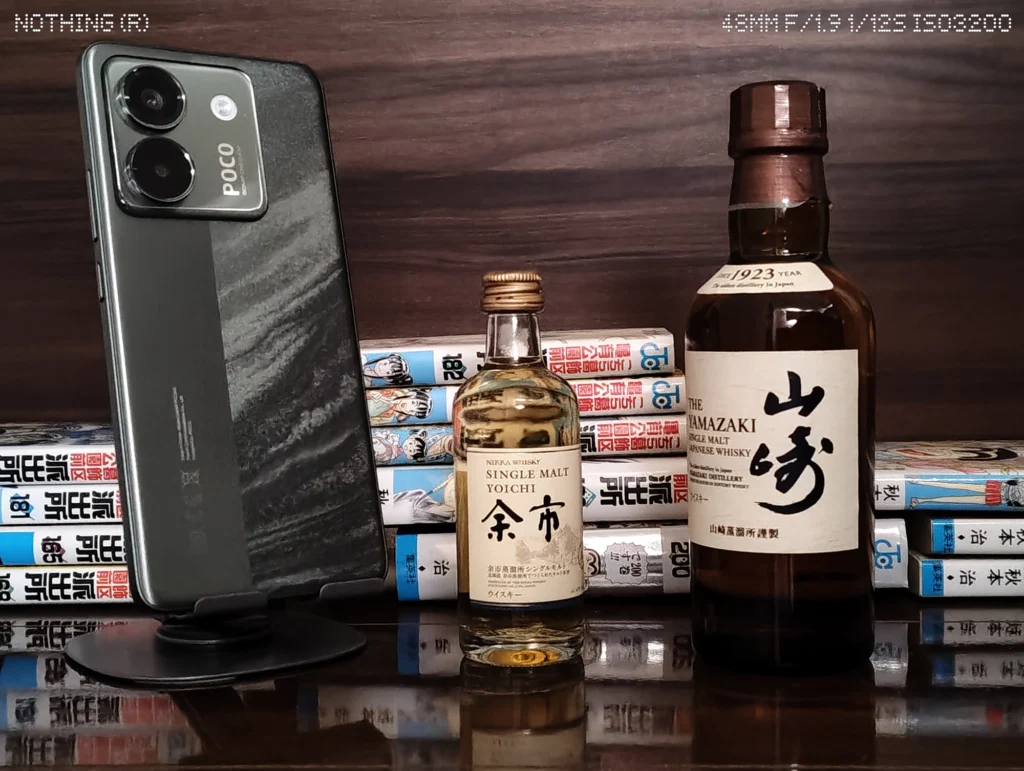Nothing Phone 4aで暗めの屋内に置いたスマホ、ウイスキーボトル、漫画本を2倍撮影したカメラ作例