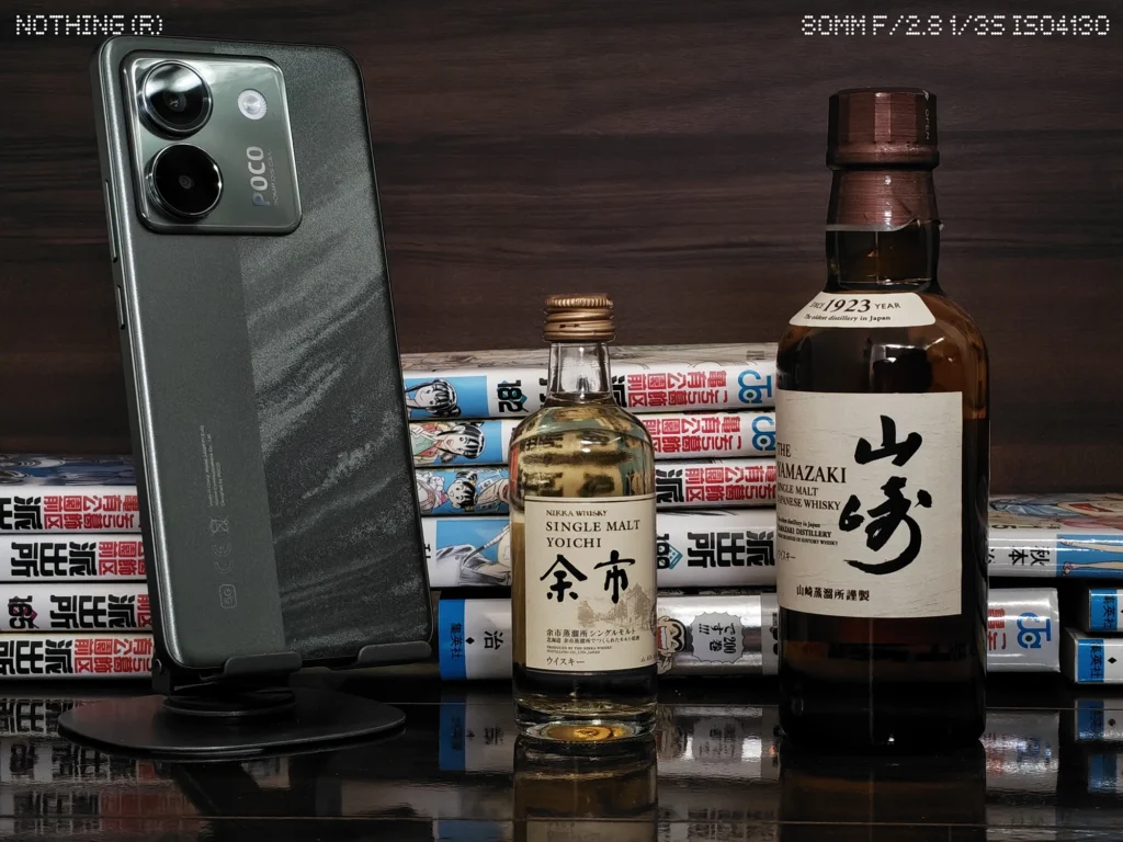 Nothing Phone 4aで暗めの屋内に並べたスマホ、ウイスキーボトル、漫画本をズーム撮影したカメラ作例