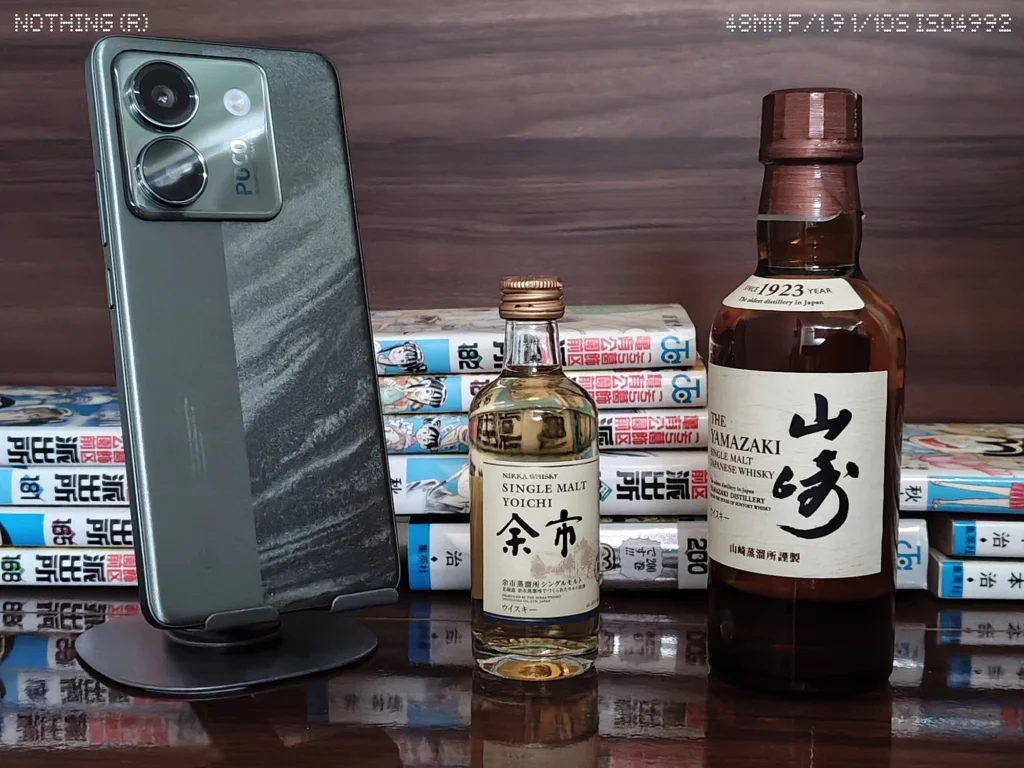 Nothing Phone 4a Proで暗めの屋内に並べたスマホ、ウイスキーボトル、漫画本を2倍撮影したカメラ作例